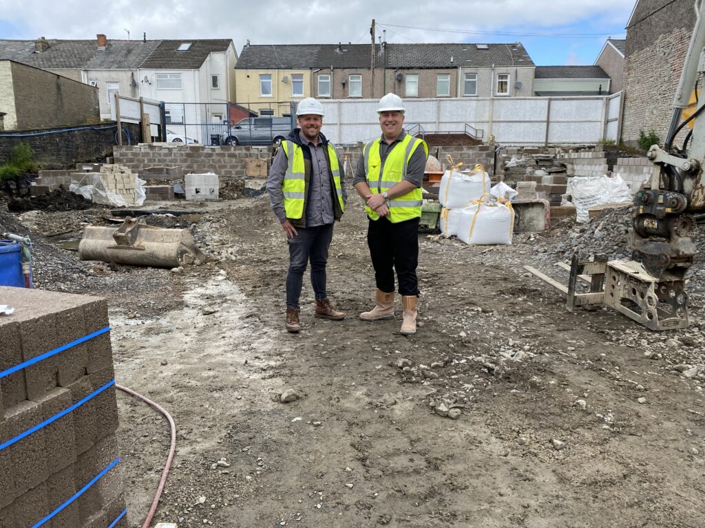 An image showing two men wearing white hard hats and yellow safety jackets stood in the centre of a building site, smiling at the camera