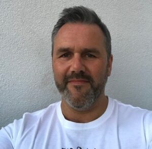 An image of a man with white skin and dark hair smiling at the camera wearing a white t shirt