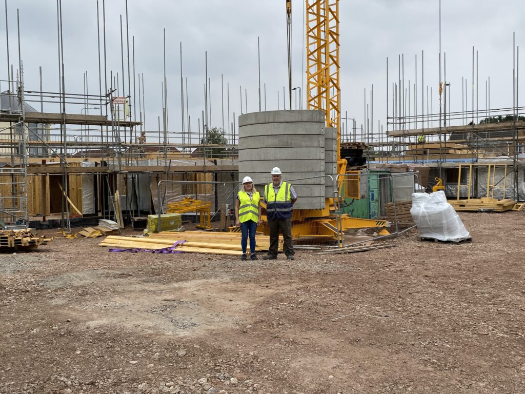 An image of a man and woman stood in the centre of a construction site smiling at the camera. They are wearing yellow safety jackets and white hard hats. The image conveys the progress made by United Welsh and partners at St Mellons in Cardiff to build temporary accommodation for families in need.