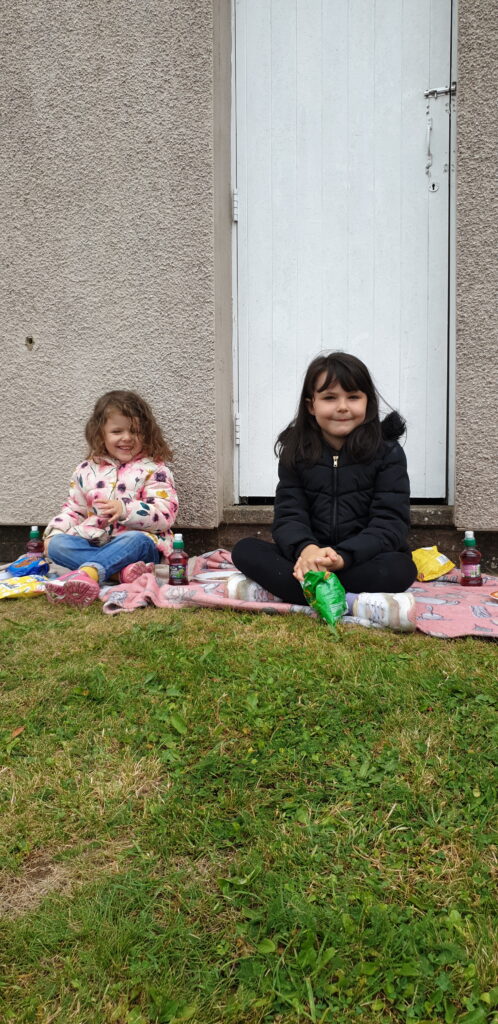 Two young girls sitting on a mat on the grass, enjoying a picnic.
