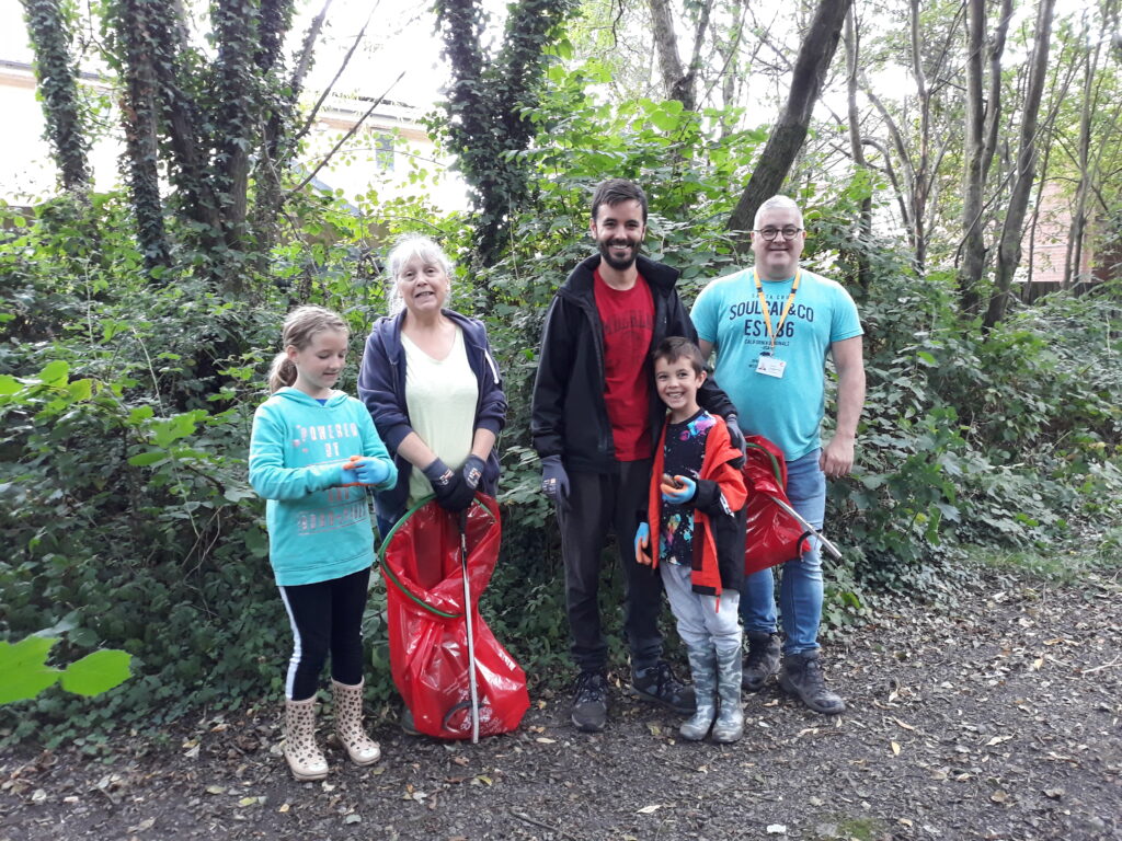 Five people standing in a forest. From L-R: A young girl with blonde hair wearing a blue hoodie and black leggings. A woman with white hair wearing a white top holding a red rubbish bag and a litter picking tool. A man wearing a red top and a black hoodie with dark hair and a beard, with his arm around his son who has dark hair, a black tshirt and a red jacket. A man with glasses wearing a blue top and jeans.