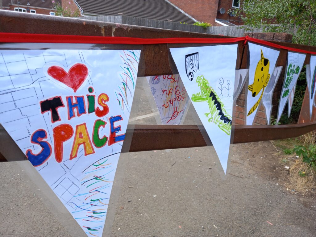 Hand-made bunting by children, tied on a fence with red ribbon. The first bunting triangle says 'love this space' in coloured pen. The second one features a drawing of a dinosaur.