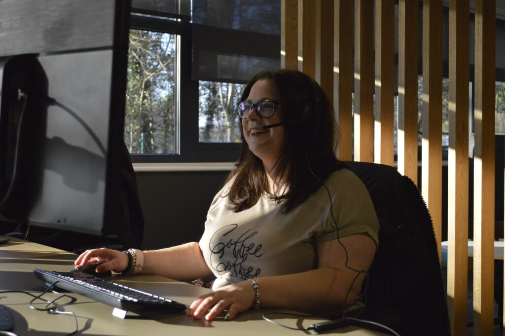 A photo of a woman sitting at a computer desk, smiling at the screen.