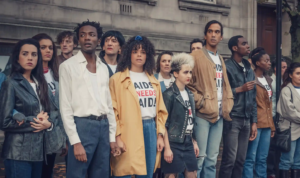 A screengrab of a scene from the Channel 4 show 'It's A Sin'. A group of people are standing at a HIV protest wearing t-shirts that say 'AIDS NEEDS AID.'