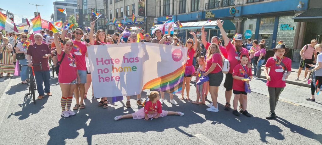A photo of a group of staff attending Pride Cymru. Everyone is wearing pink t-shirts with a rainbow on the front and holding a banner that says 'hate has no home here'.
