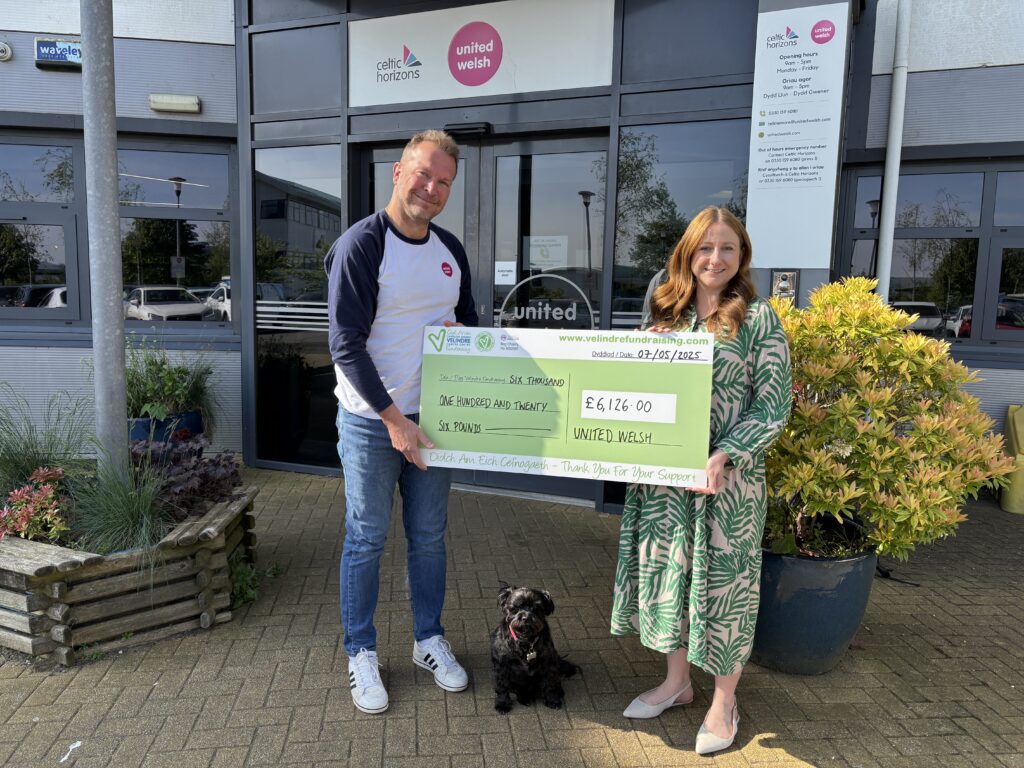 A photo of United Welsh Group CEO Richard Mann and Fundraising Manager Emma Prince from Velindre. They are standing outside United Welsh's office building, holding a large Velindre check.