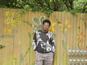 Photo of Timi in the garden of Oak House. Timi is standing in front of a fence that has a mural with yellow and green flowers on it.