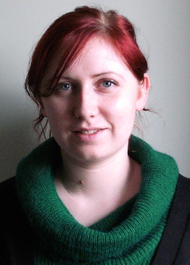 A photo of a woman with dark red hair tied back, wearing a green turtle neck jumper.