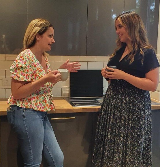A photo of two women chatting in a kitchen, holding mugs.