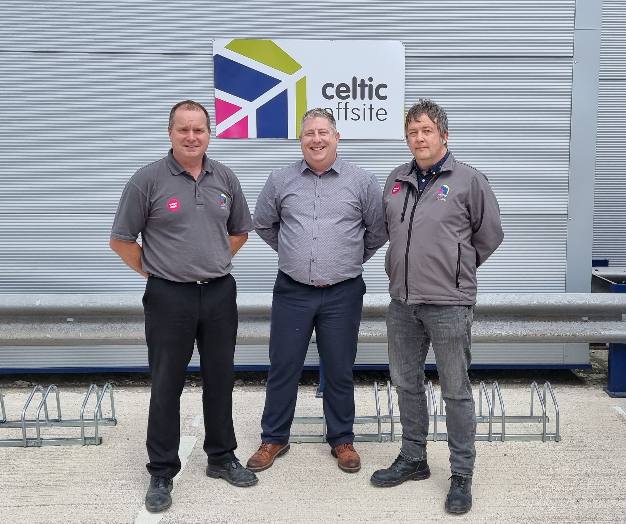 Three men stood facing the camera, wearing grey shirts and fleeces in front of a grey metal panel wall