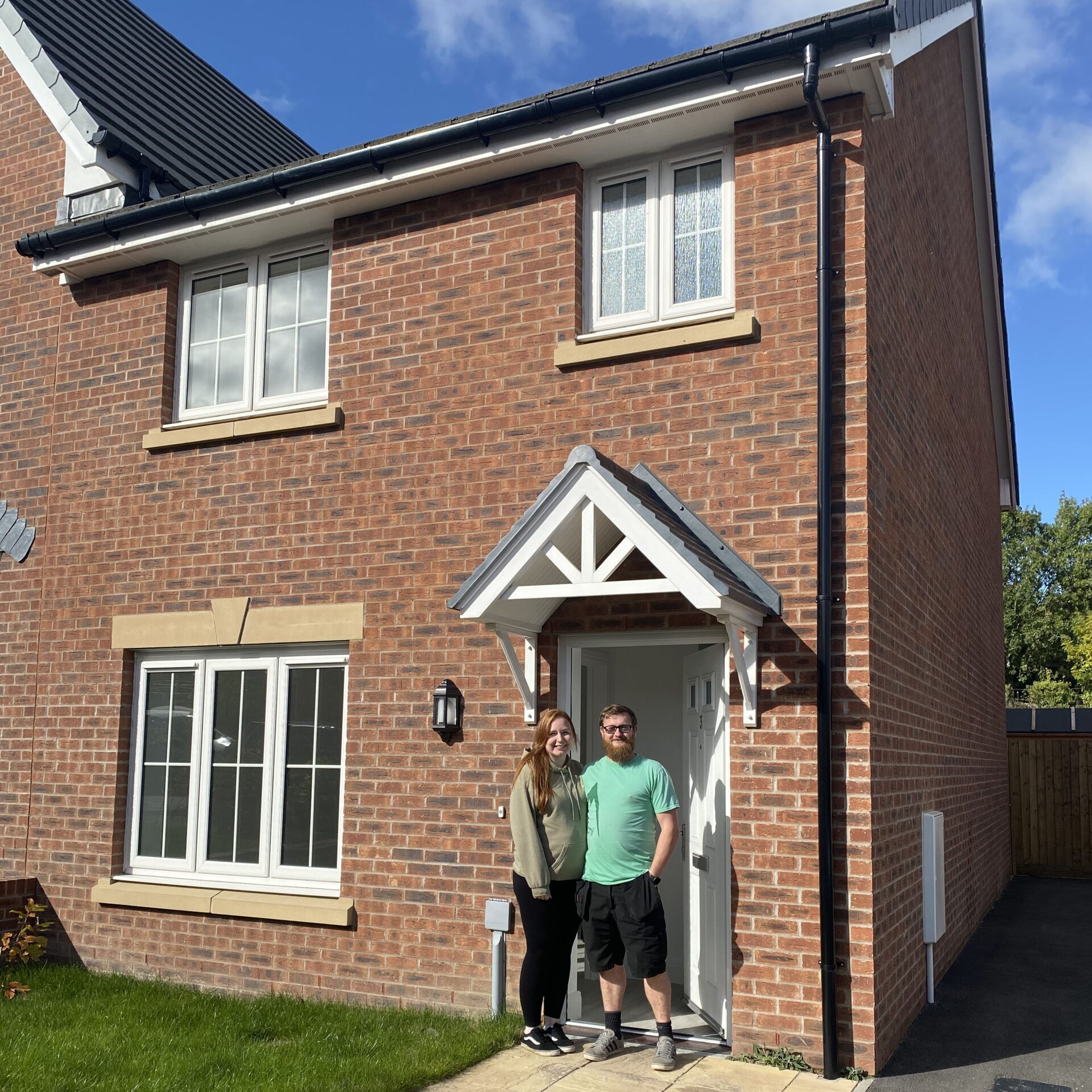An image of a hapy couple smiling in the doorway of their new home. The home has red brick render and a white gable above the front door