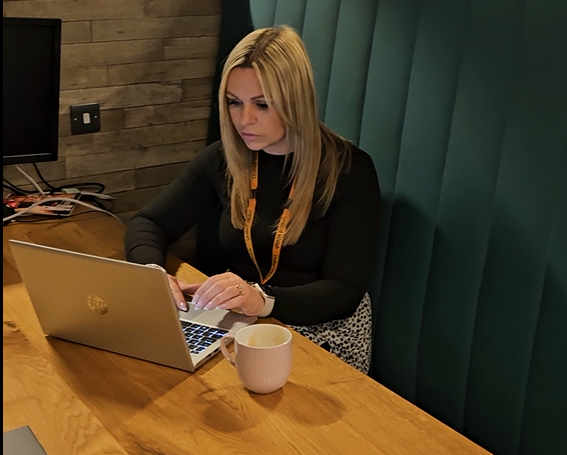 Woman with blond hair sitting at a desk working at a laptop with a cup of coffee next to her.